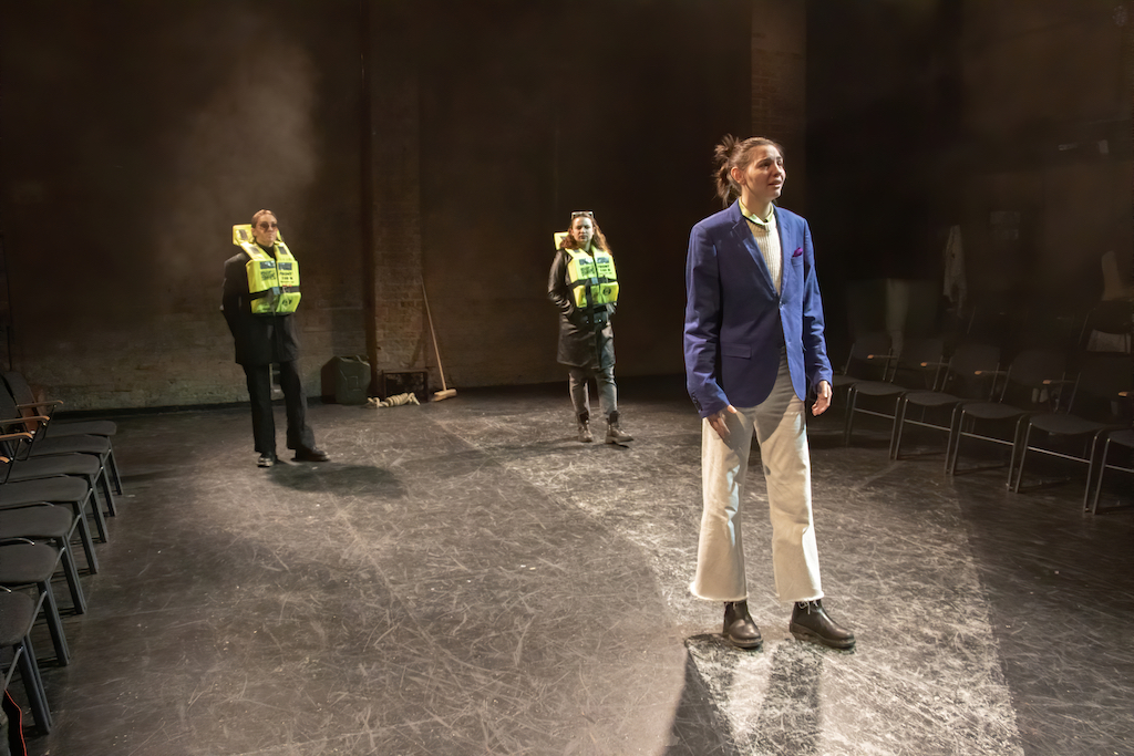 An actor, wearing a blue blazer and white trousers, stands downstage while two additional actors wearing neon yellow life jackets and sunglasses stand behind them.